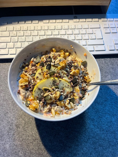 Karotten-Apfel-Müsli in weißer Porzellan-Schale auf einem grauen Tisch neben einer Computertastatur