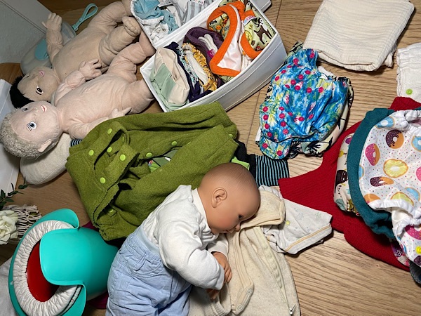 Auf einem Holztisch liegen verschiedene Babypuppen, bunte Stoffwindeln, spezielle Windelfrei-Bekleidung und Abhaltetöpfchen.