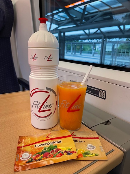 Fitline Trinkflasche, Becher mit Getränk Powercocktail auf Tisch im Zug, mit Blick durchs Fenster auf einen Bahnhof