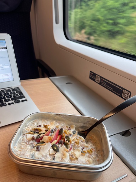 Müsli mit Apfelscheiben und Nüssen in rechteckiger Edelstahldose vor geöffnetem Laptop auf Tisch im ICE