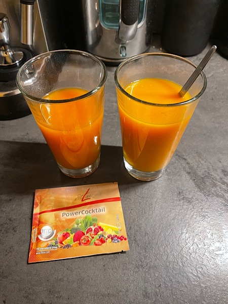 Zwei große Gläser ohne Aufdruck mit orangefarbenem Inhalt, der Powercocktail genannt wird, auf einer grauen Platte.