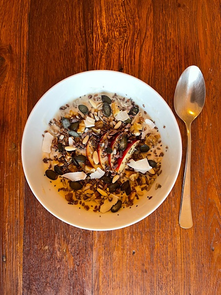 Müsli in weißer Schale von oben fotografiert auf dunkelbraunem Holztisch mit silbernem Löffel rechts daneben