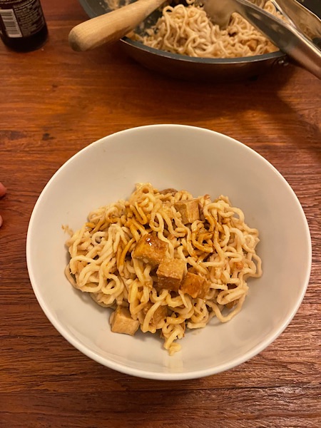 Mie-Nudeln mit Tofu und Erdnuss-Sauce in weißer Porzellanschale auf dunkelbraunem Tisch von oben fotografiert