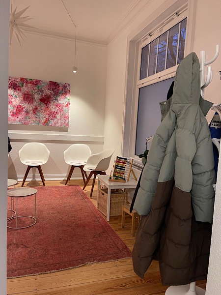 Wartezimmer beim Arzt mit hohen Decken, weißen Wänden, Holzboden, rotem Teppich, weißen Stühlen, Garderrobe mit Winterjacken im Vordergrund