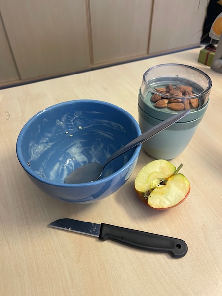 leere blaue Müslischale, halber aufgeschnittener Apfel, Obstmesser und Joghurtaufbewahrungsdose mit Mandeln oben drin